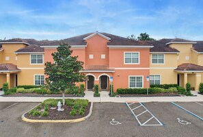A multi-story residential building with a central entrance, a parking lot with handicapped parking spaces, and a small tree in the foreground. - Spacious 4BR Retreat | Pool | Disney & Universal (Kissimmee)