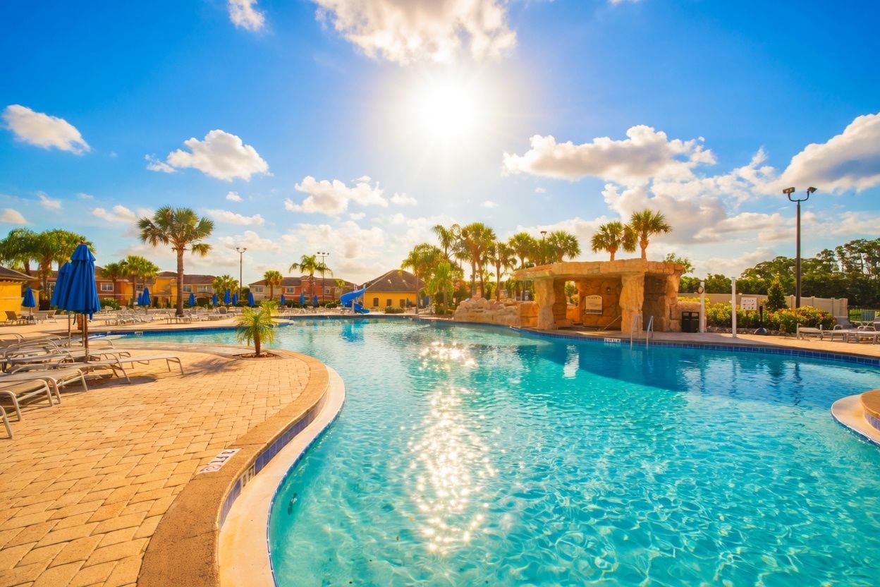 A swimming pool with lounge chairs and umbrellas, surrounded by palm trees and buildings.