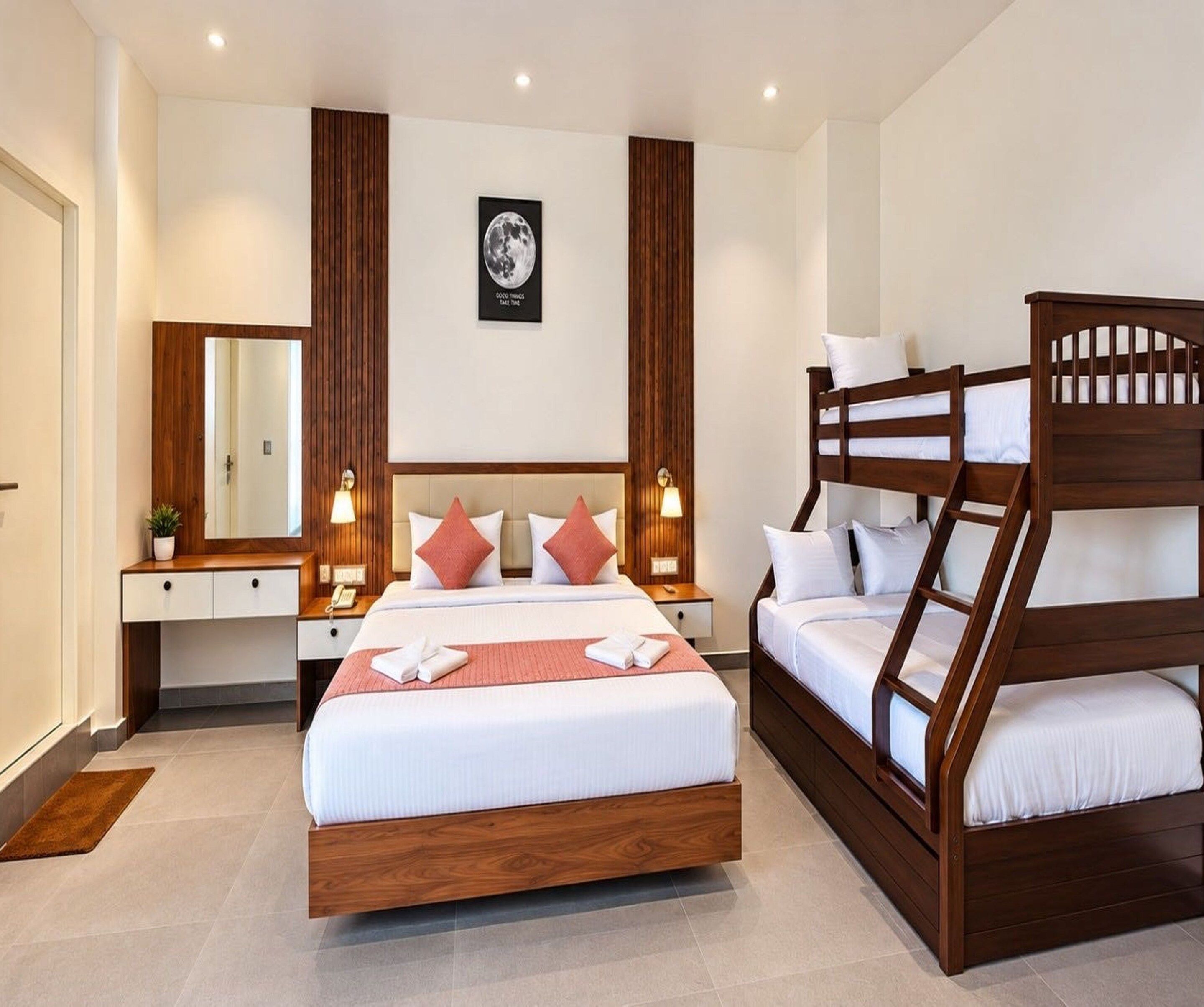 A hotel room with a large bed, a bunk bed, a bedside table with a lamp, a mirror, and a framed picture on the wall.