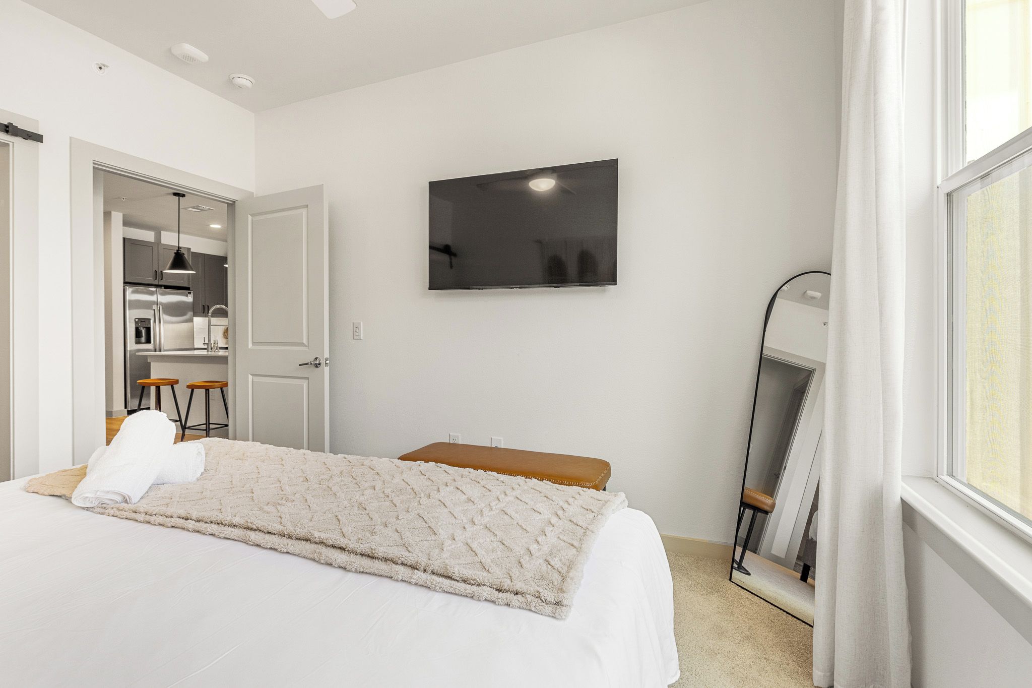 A bedroom with a bed, a flat-screen TV, a mirror, and a door leading to a kitchenette.