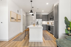 A modern kitchen with a central island, grey cabinetry, stainless steel appliances, and a bar stool. - Luxury 1 Bed with Walkout Garden MER 9115 (San Marcos)
