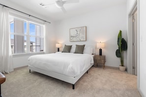 A bedroom with a large bed, two bedside tables, a ceiling fan, a window with curtains, a wall-mounted artwork, and a potted plant. - Modern 2BR 2BA | Open Layout  MER 1307 (San Marcos)