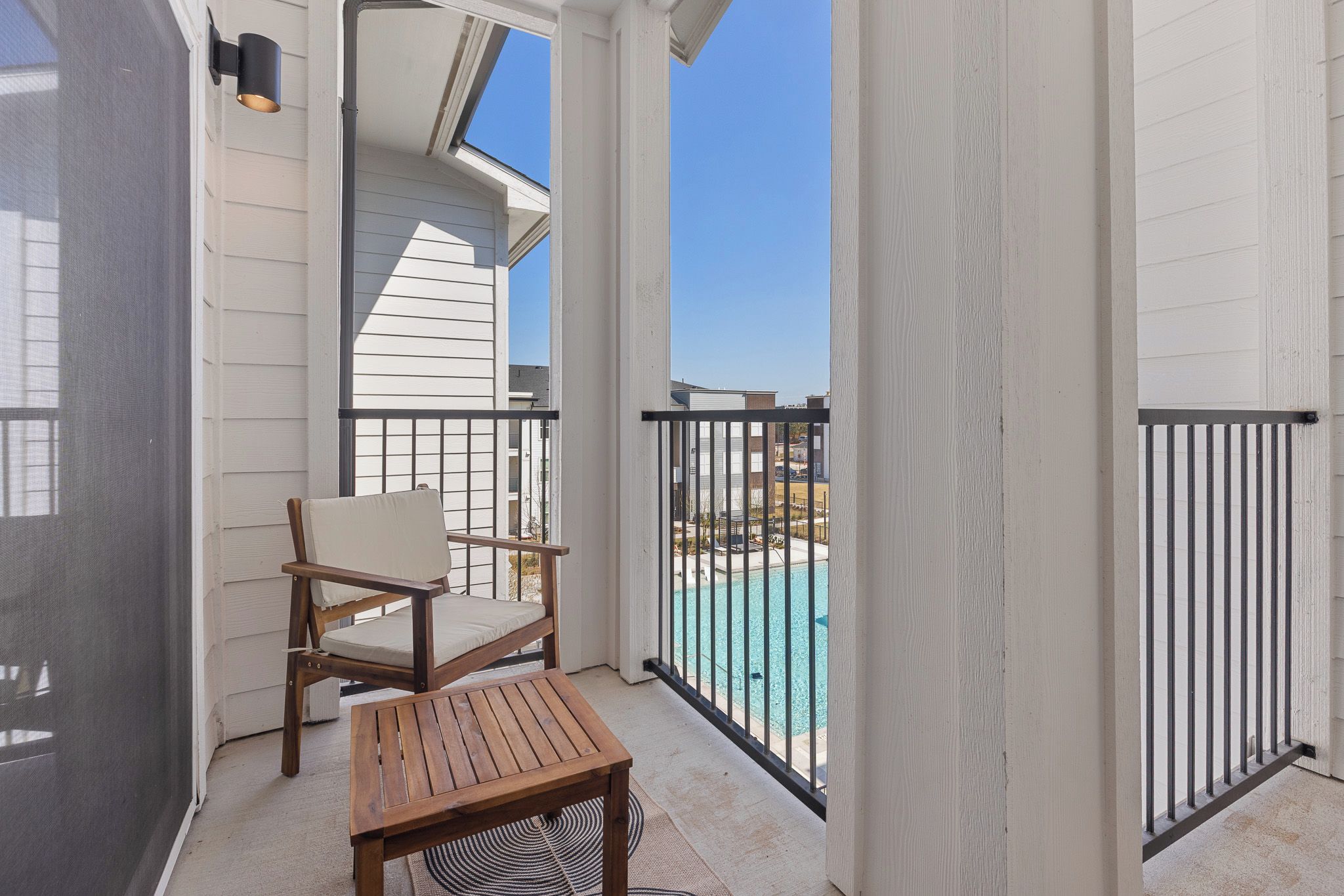 A balcony with a wooden chair and table, a pool in the background, and a clear sky.