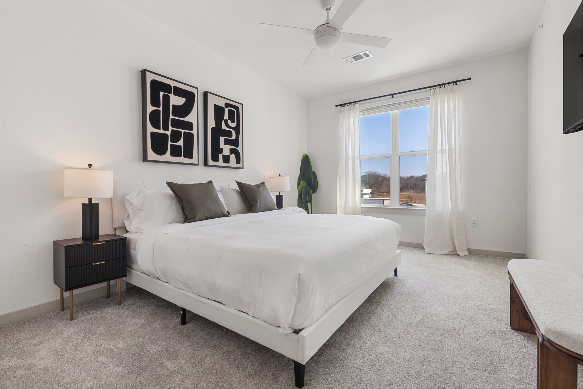 A bedroom with a bed, bedside tables, a ceiling fan, a window with curtains, and artwork on the wall.