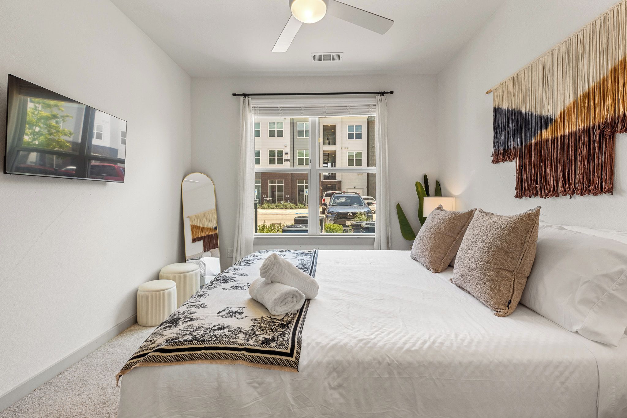 A hotel room with a bed, a large window, a ceiling fan, a mirror, and a wall-mounted tapestry.