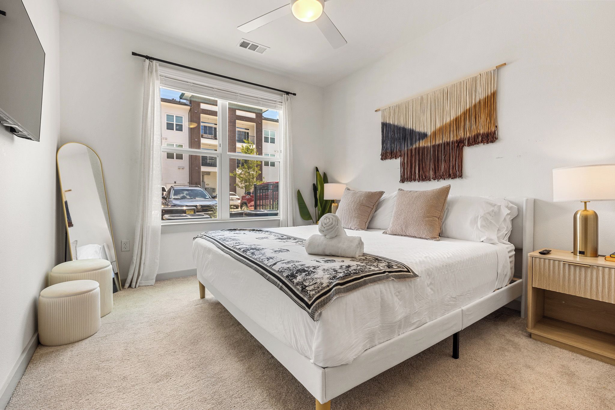 A hotel room with a bed, bedside table, lamp, and a view of the outside.