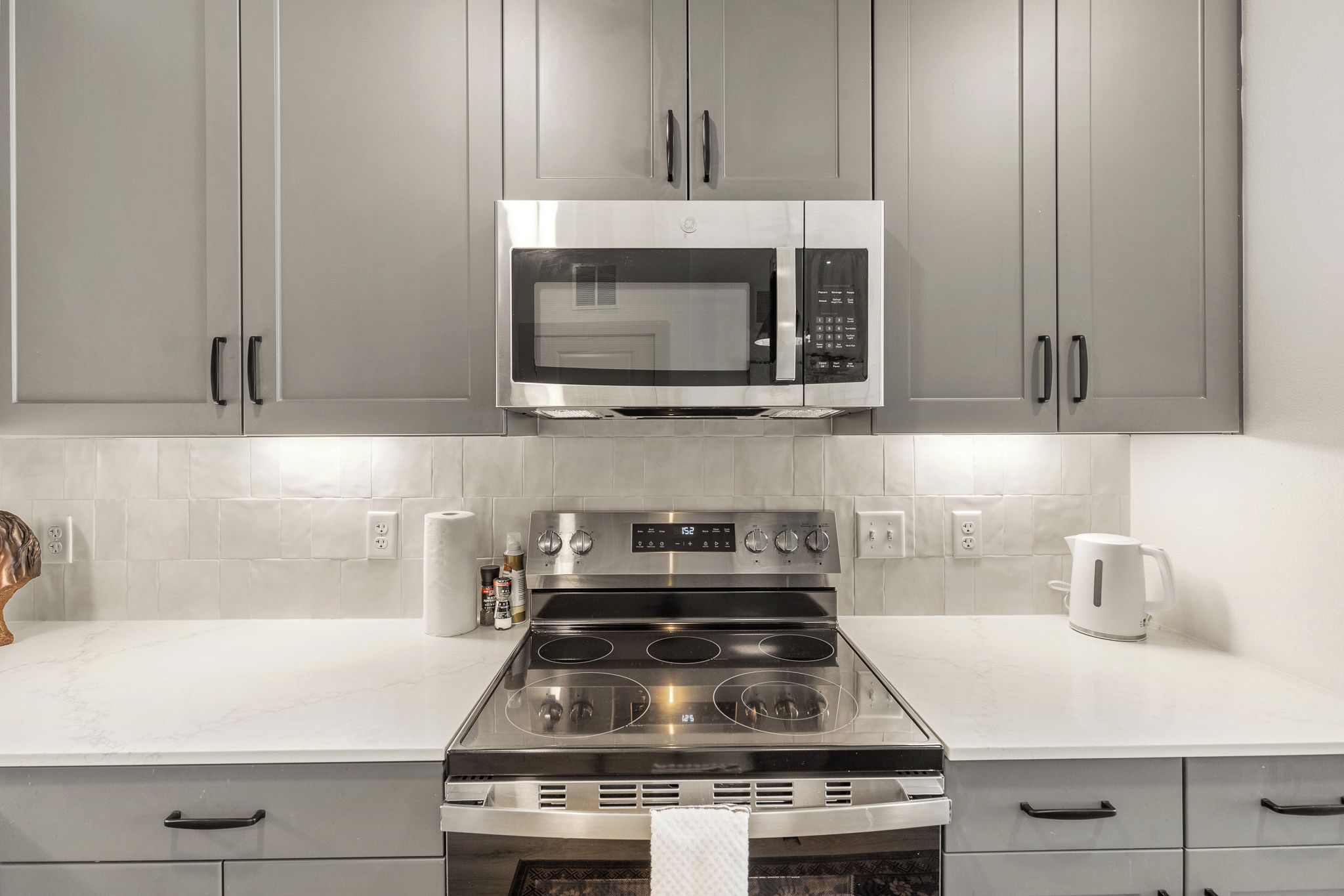A kitchen with grey cabinets, a stainless steel microwave, a gas stove, and a white countertop.