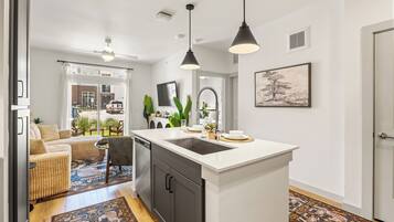 A modern kitchen with a central island, a dining area with a sofa and chairs, and a living room with a TV and plants.
