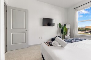 A hotel room with a bed, a flat-screen TV, a plant, and a window with a view of the outside. - Stylish 1BR Stay | Pool + Luxe Amenities MER 3106 (San Marcos)