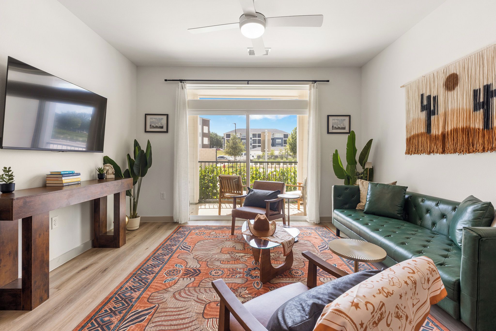 A modern living room with a sofa, coffee table, and chairs, a flat-screen TV mounted on the wall, and a balcony with a view of buildings.