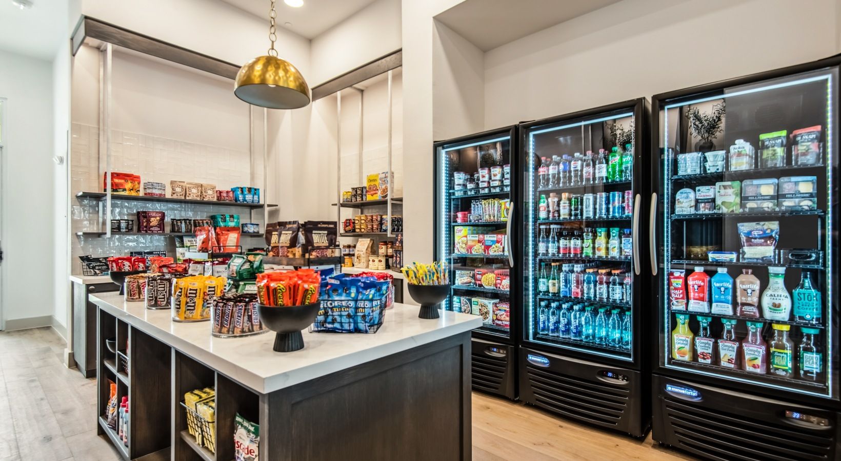 A well-stocked beverage refrigerator with various drinks, a central counter with snacks and packaged goods, and a hanging light fixture.