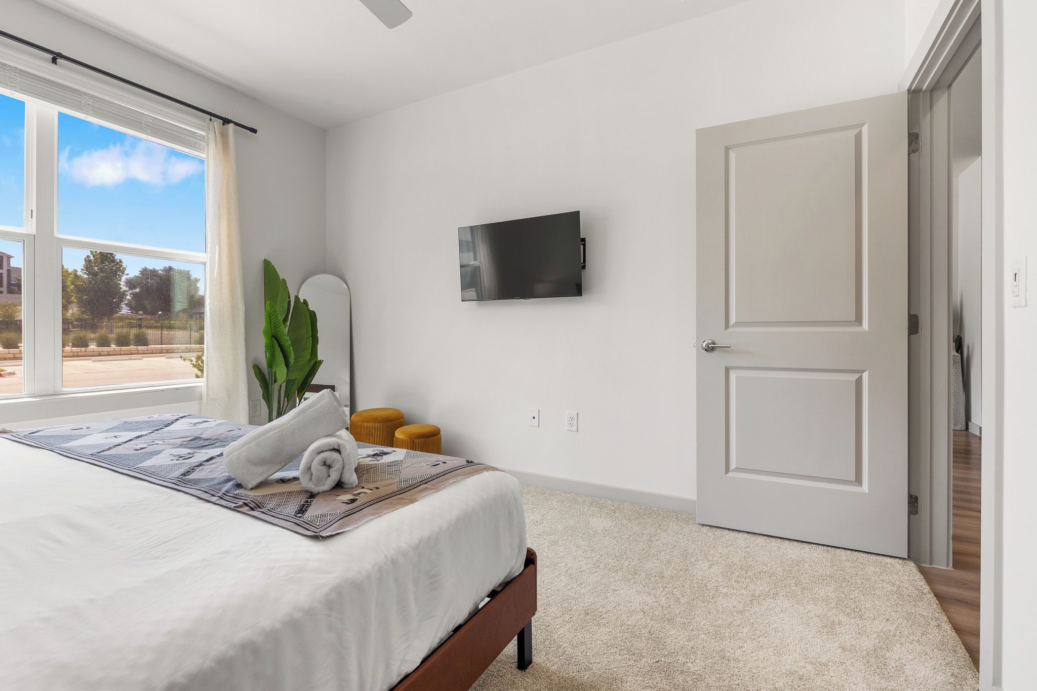 A bedroom with a bed, a television mounted on the wall, a door, and a window with a view of the outside.