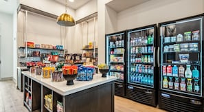 Een goed gevulde drankkoelkast met diverse dranken, een centraal gelegen toonbank met snacks en verpakte producten, en een hangende lamp.