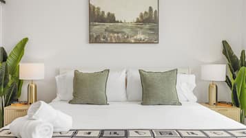Een hotelkamer met een bed, twee groene kussens, wit beddengoed, een schilderij boven het bed en twee nachtkastjes met lampen en boeken.