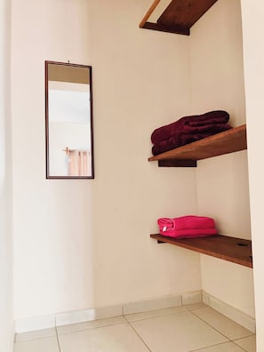 Eine Ecke eines Zimmers mit einem Holzregal, auf dem ein gefaltetes Handtuch liegt und einem Spiegel an der Wand.