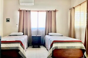 Ein Hotelzimmer mit zwei Betten, einem kleinen Nachttisch, einem Fenster mit Vorhängen und einer Wand-Klimaanlage.