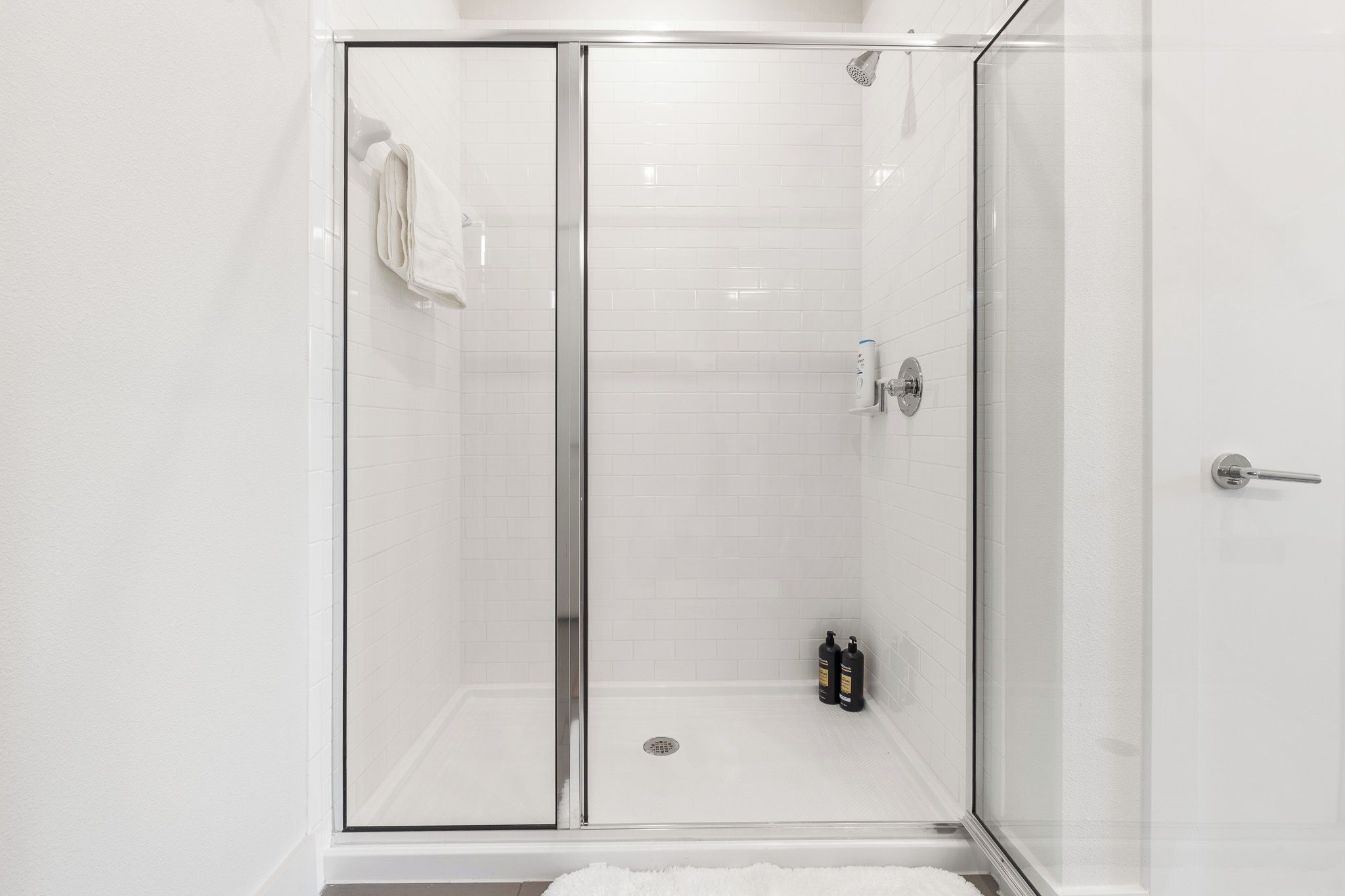 Une cabine de douche fermée par des parois en verre, avec un mur recouvert de carreaux blancs, une serviette accrochée à la porte et deux bouteilles posées sur le sol.