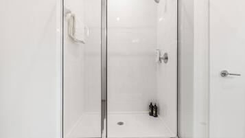 Une cabine de douche fermée par des parois en verre, avec un mur recouvert de carreaux blancs, une serviette accrochée à la porte et deux bouteilles posées sur le sol.