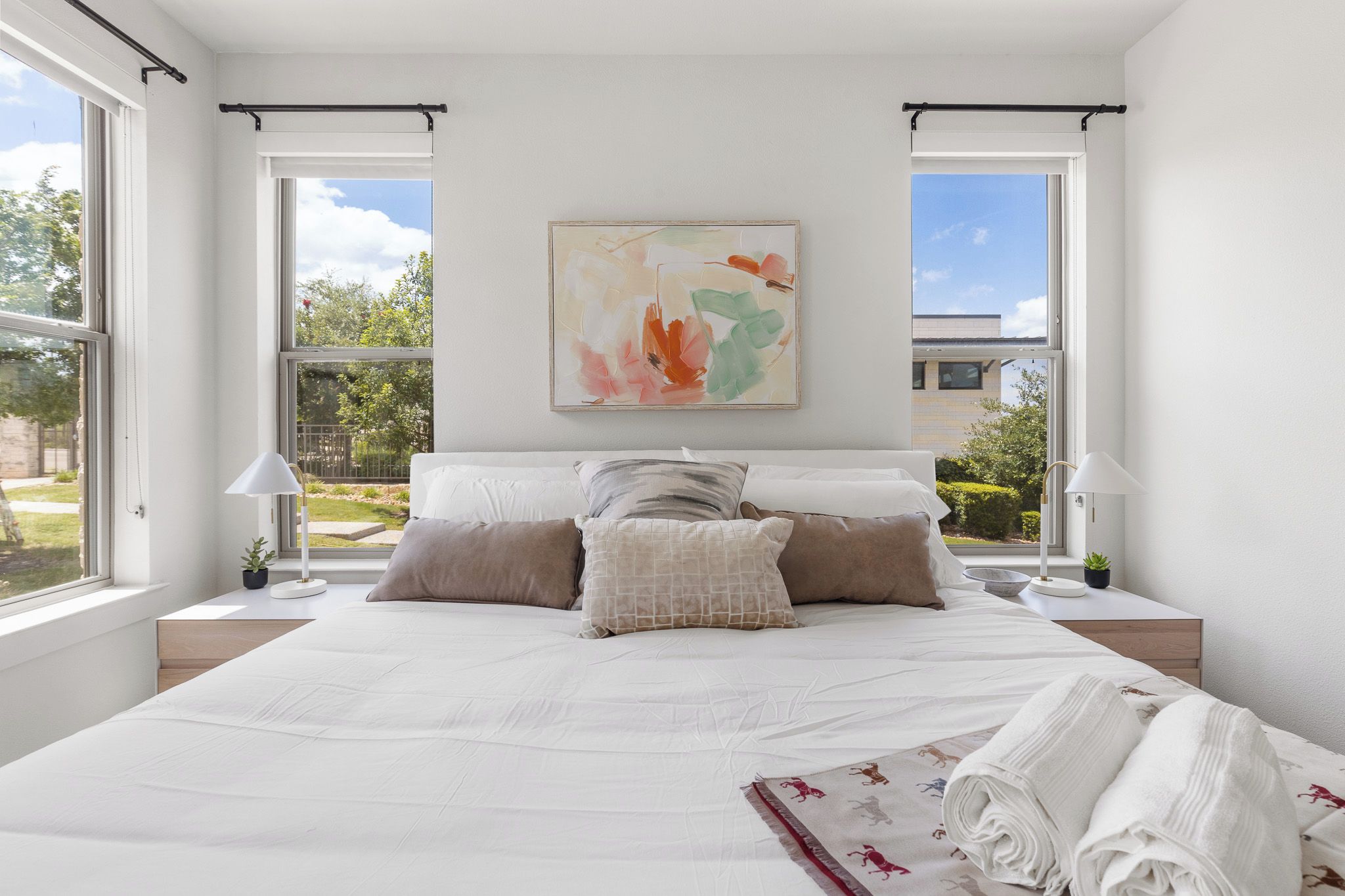 A modern bedroom with a large bed, two bedside tables, a nightstand, a painting, and large windows.