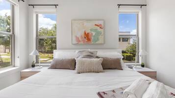 A modern bedroom with a large bed, two bedside tables, a nightstand, a painting, and large windows.