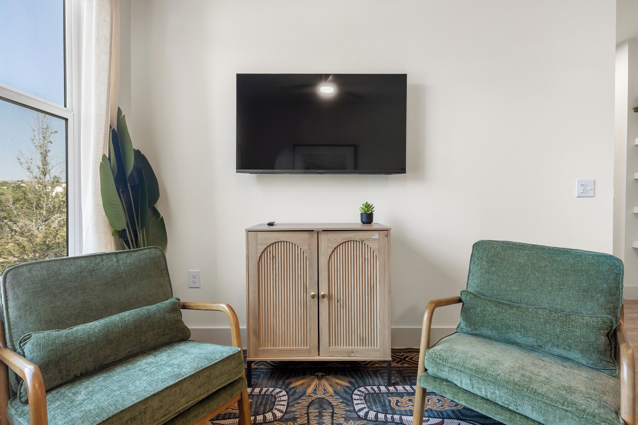 A room with two green upholstered chairs, a wooden cabinet with a flat-screen TV mounted above, and a patterned carpet.