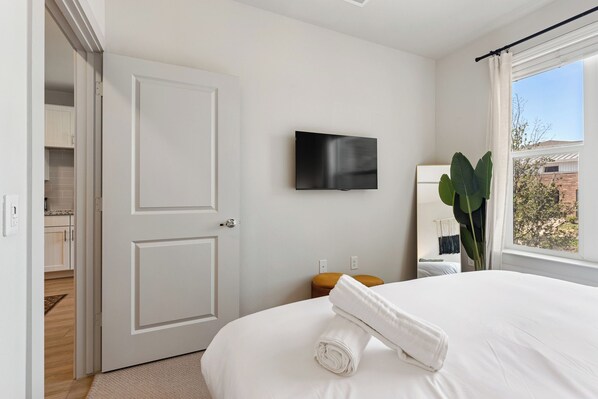 A hotel room with a bed, a flat-screen TV mounted on the wall, a window with a view of the outside, and a door leading to a bathroom.