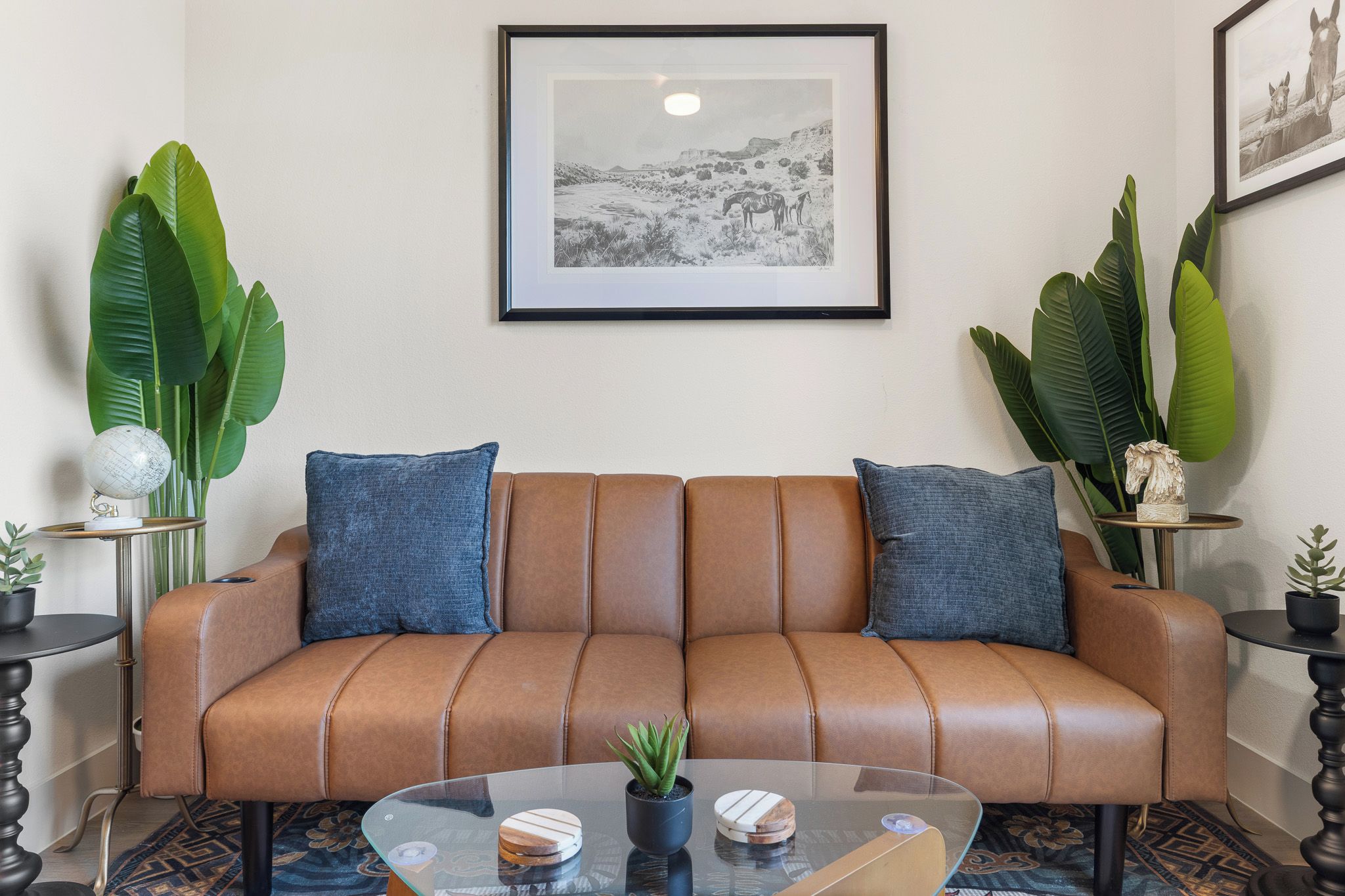 A brown leather sofa with blue cushions, a glass coffee table with decorative items, and framed artwork on the wall.