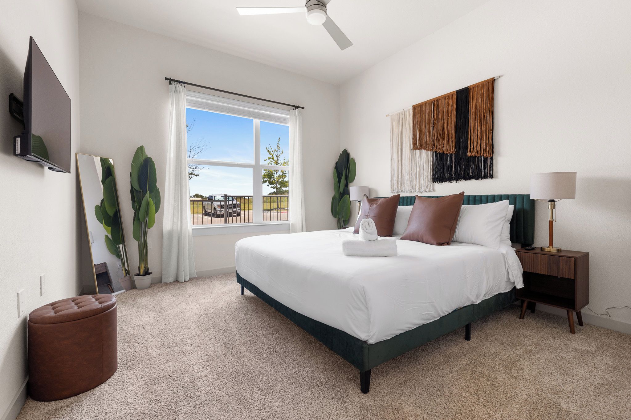 A bedroom with a bed, a nightstand, a lamp, a TV, and a window with a view of the outside.