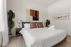A bedroom with a large bed, a nightstand, a lamp, a wall hanging, and a painting. - Cozy Collin Square 1BR | Relax + Work CSA 5107 (McKinney)