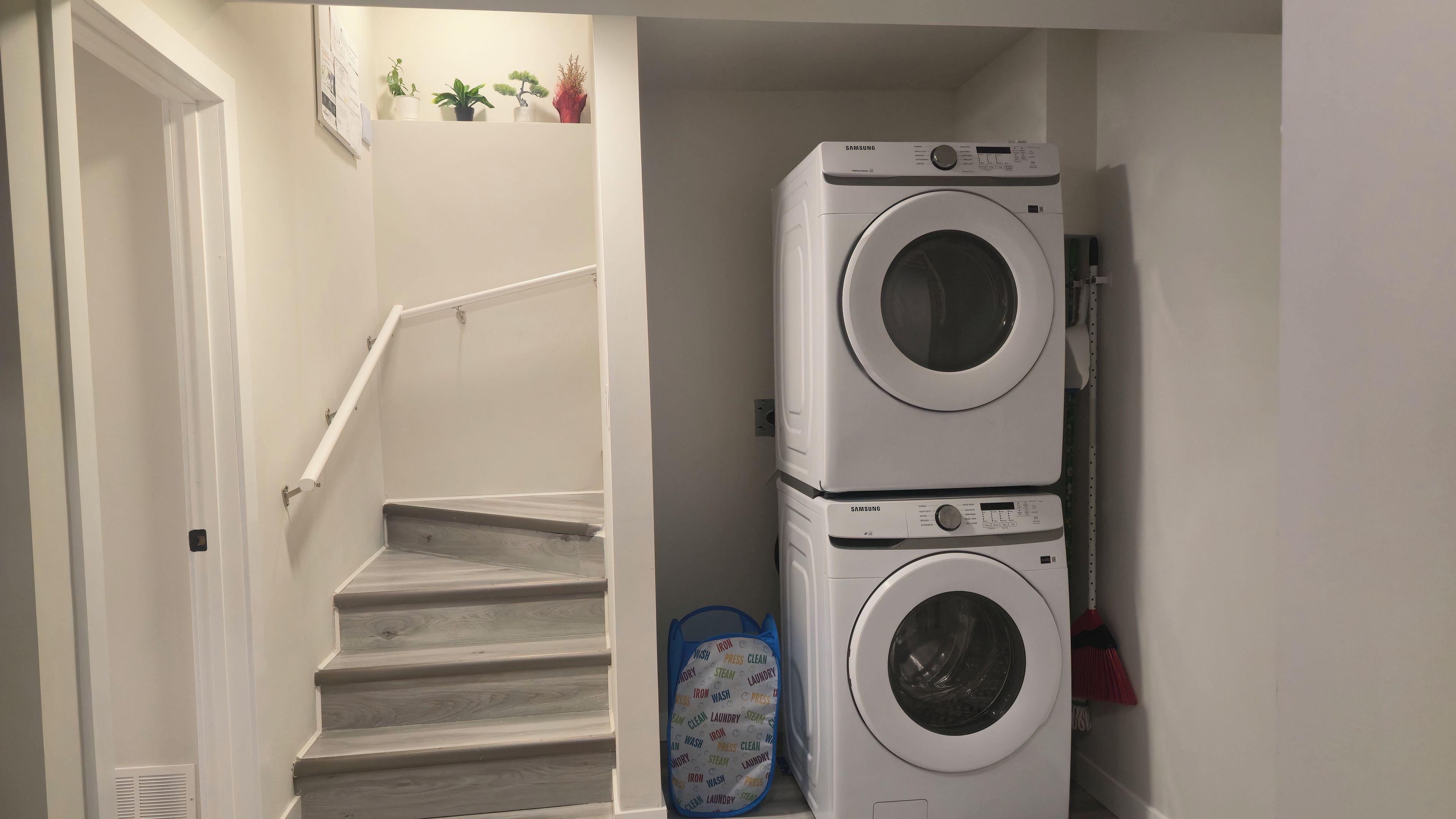 A laundry room with two stacked washing machines, a stairway, and a blue laundry basket with labels.