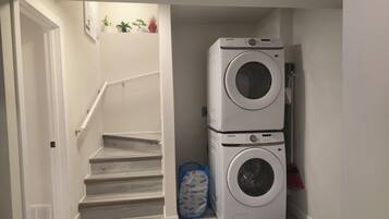 A laundry room with two stacked washing machines, a stairway, and a blue laundry basket with labels.