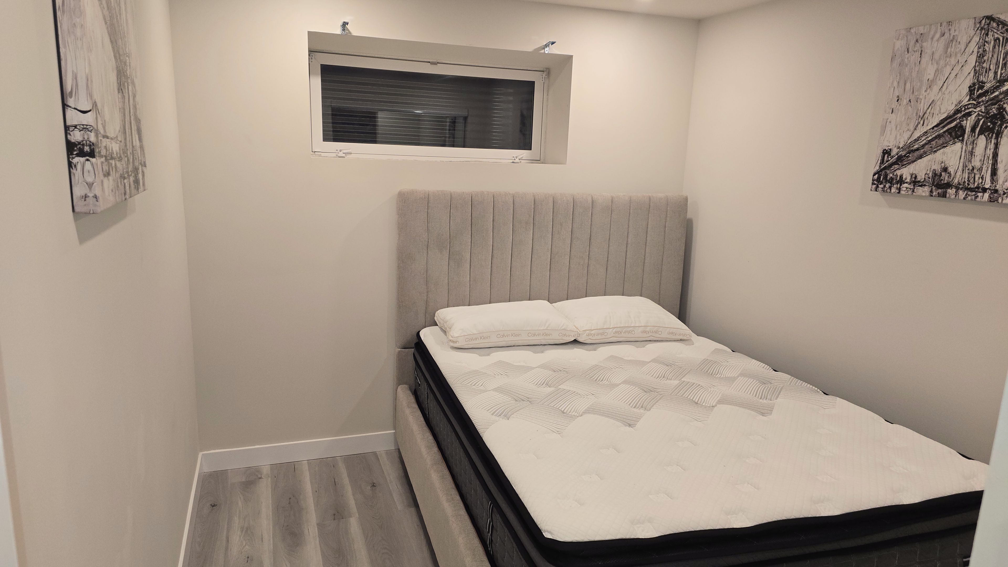 A small, neatly arranged bedroom with a single bed, a window, and artwork on the walls.