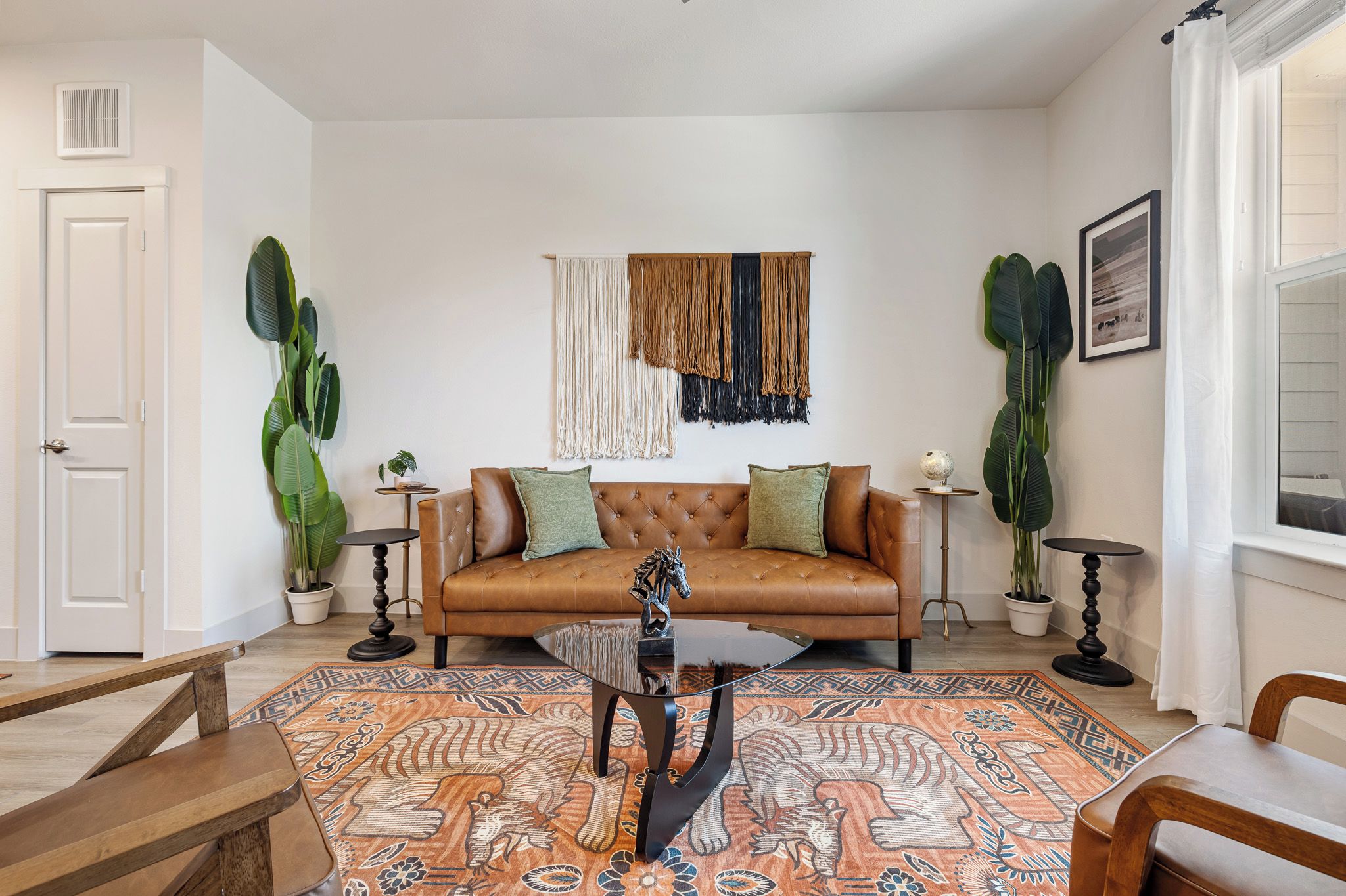A living room with a brown leather sofa, a glass coffee table, and patterned rugs. There are two potted plants, a framed picture on the wall, and a white door.