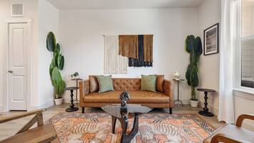 A living room with a brown leather sofa, a glass coffee table, and patterned rugs. There are two potted plants, a framed picture on the wall, and a white door.