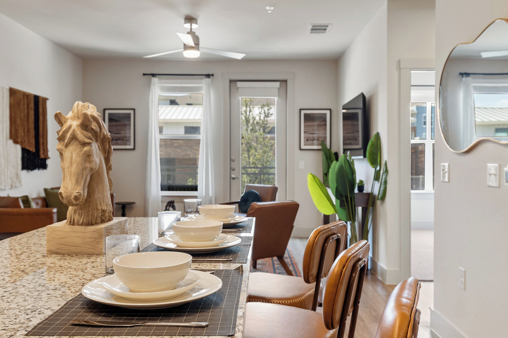 A dining area with a long table set for four, a ceiling fan, and a view through the window.