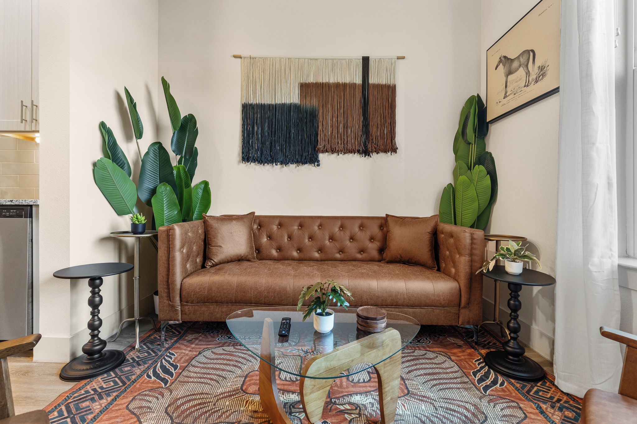 A living room with a brown leather sofa, a glass coffee table, and two side tables with potted plants.