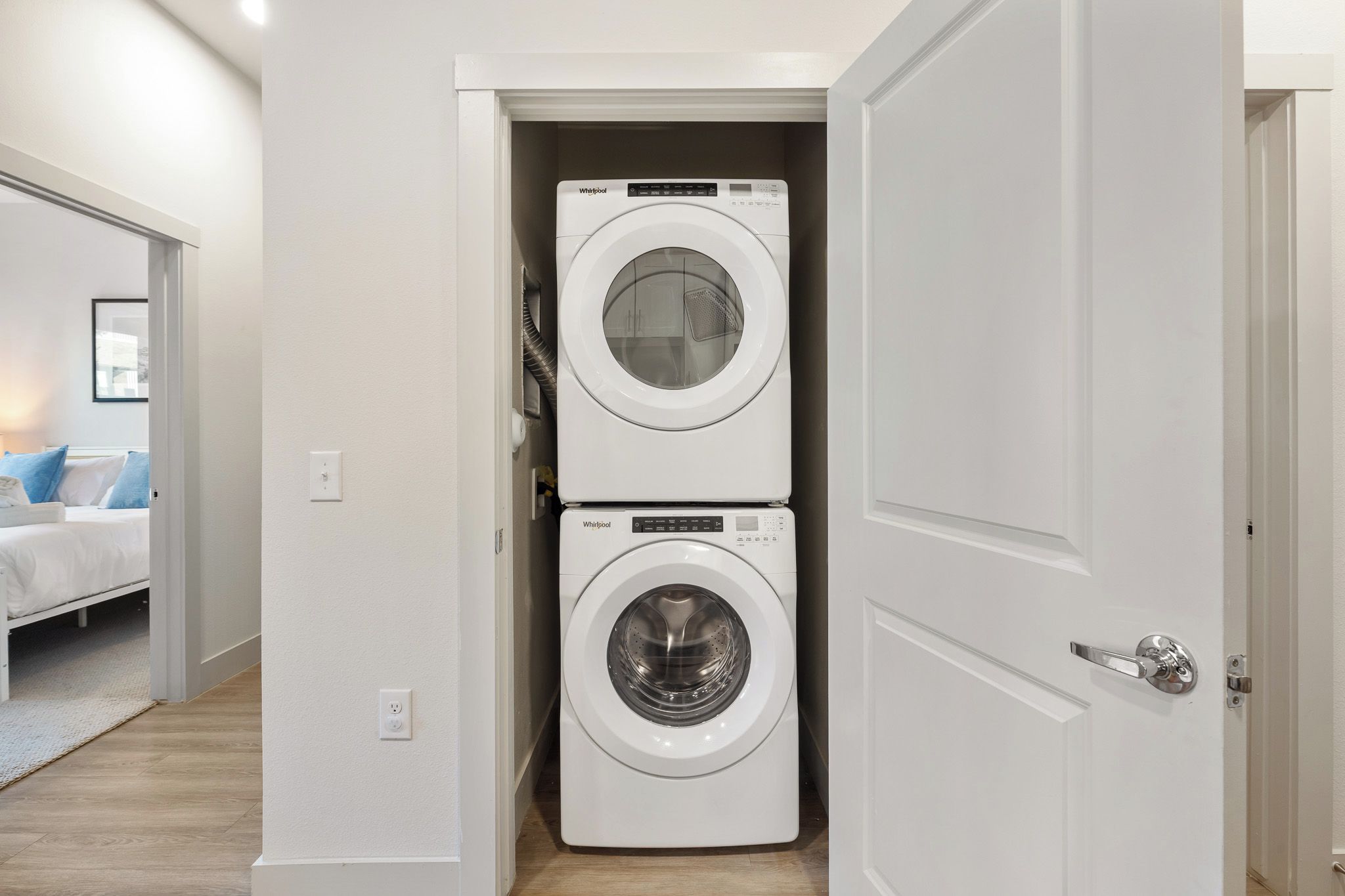 A laundry room with two stacked Whirlpool washing machines, a bed with blue pillows, and a door leading to another room.