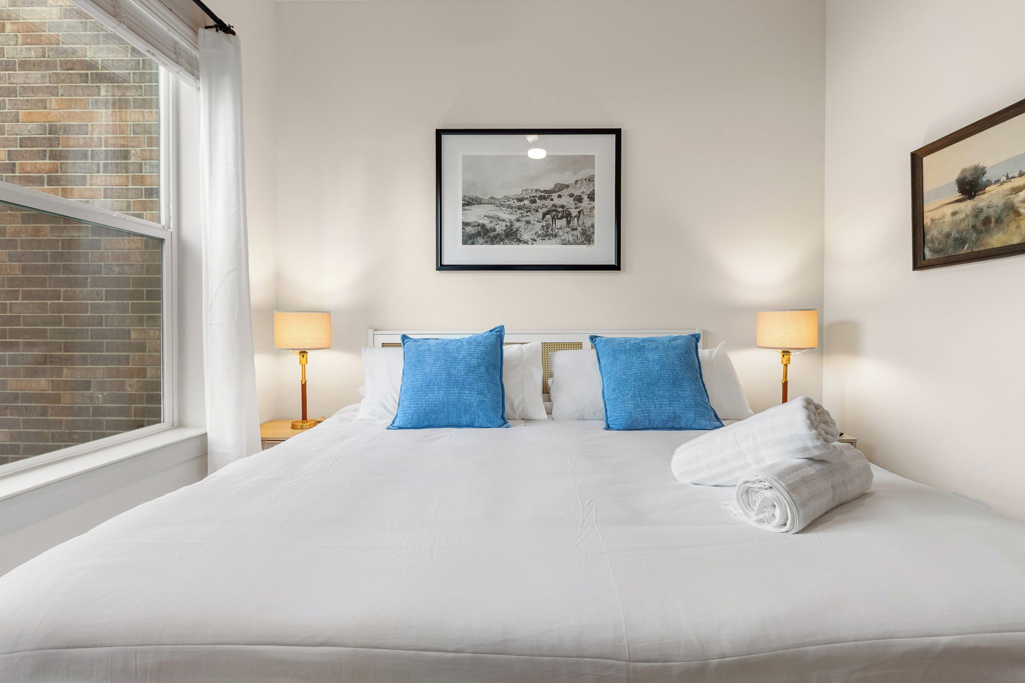 A hotel room with a large bed, two blue pillows, a white blanket, a rolled towel, a framed picture on the wall, and a window with a view of a brick wall.