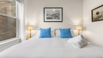 A hotel room with a large bed, two blue pillows, a white blanket, a rolled towel, a framed picture on the wall, and a window with a view of a brick wall.
