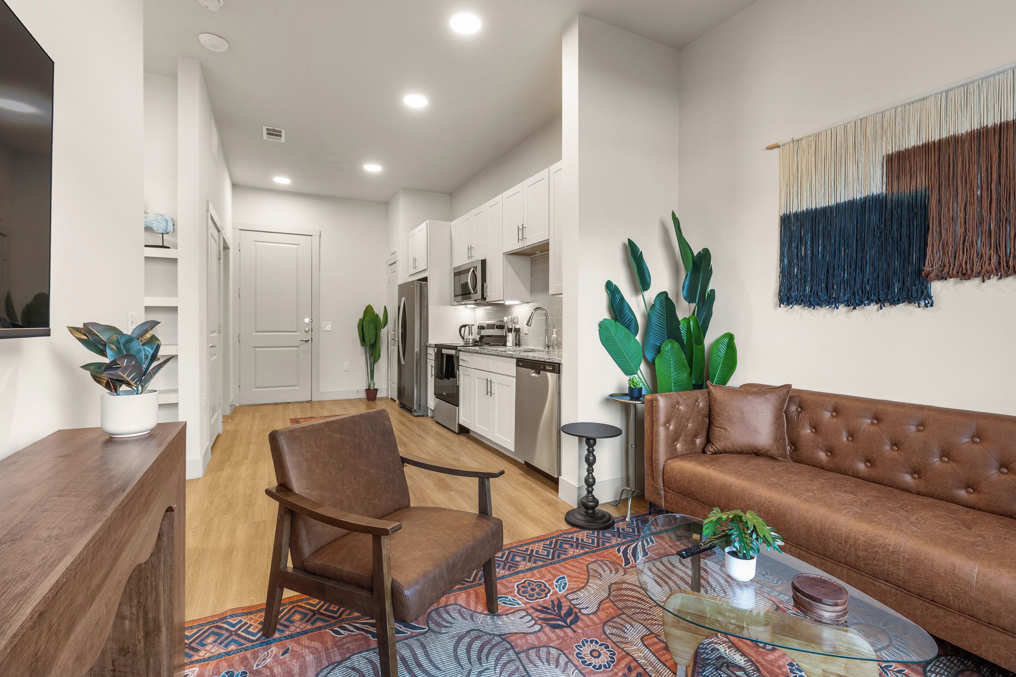 A modern living room with a brown leather sofa, a wooden chair, a glass coffee table, and a patterned rug.