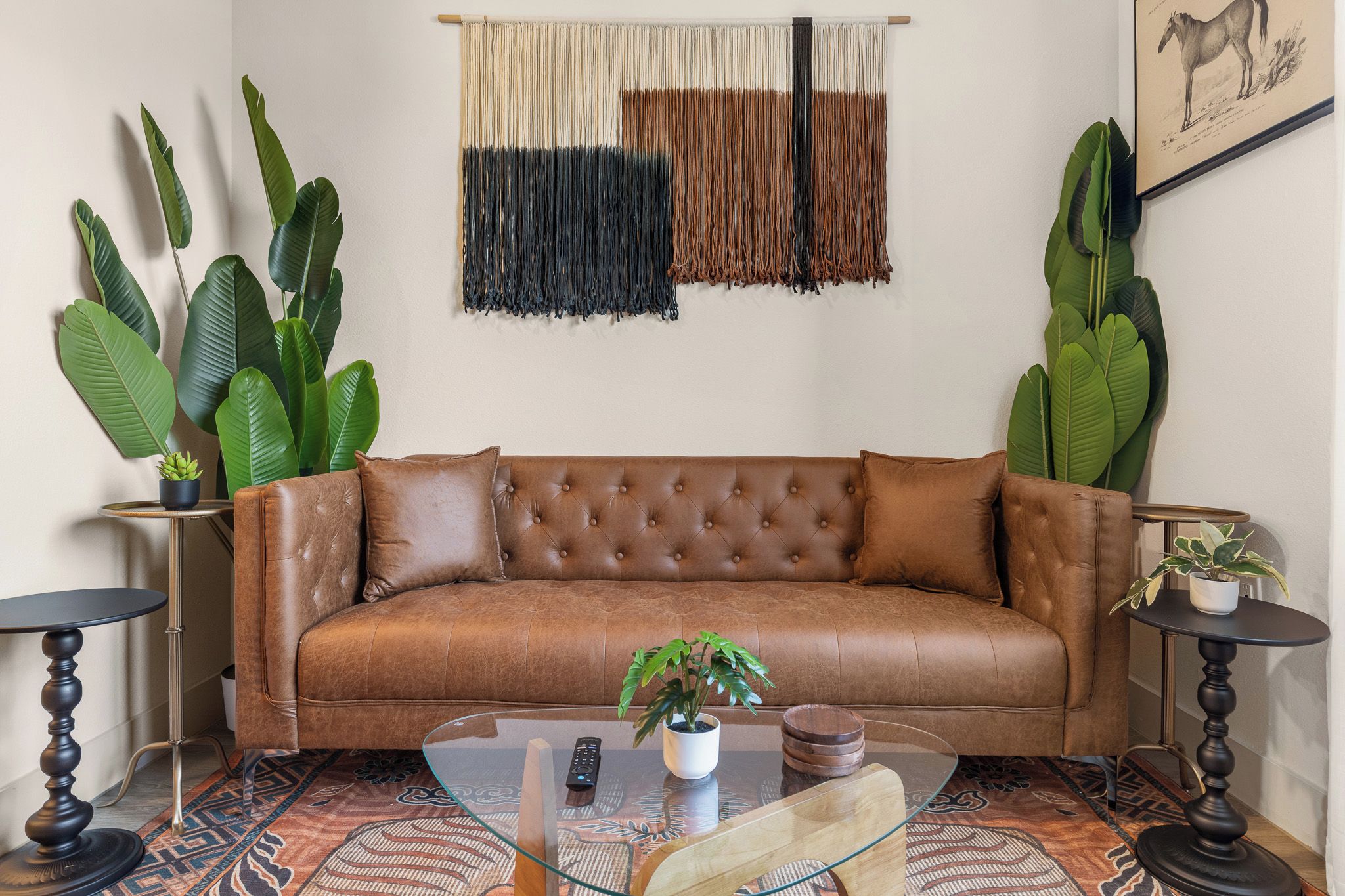 A brown leather sofa with a tufted backrest, flanked by two round side tables with dark wooden bases and black tops. A glass coffee table with a wooden top is in front of the sofa, and a small potted plant is on the right side table.