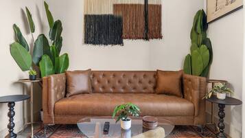 A brown leather sofa with a tufted backrest, flanked by two round side tables with dark wooden bases and black tops. A glass coffee table with a wooden top is in front of the sofa, and a small potted plant is on the right side table.