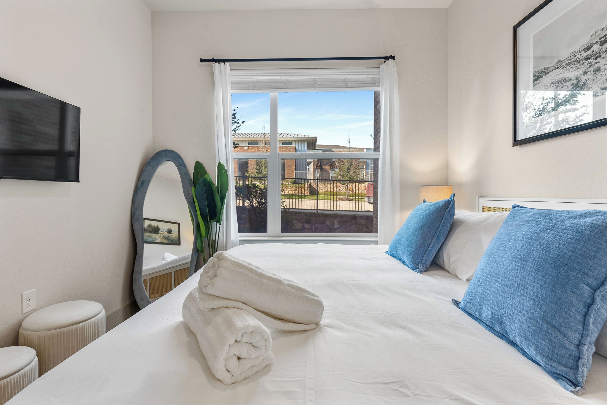 A hotel room with a bed, a window with a view of buildings, a mirror, and a plant.