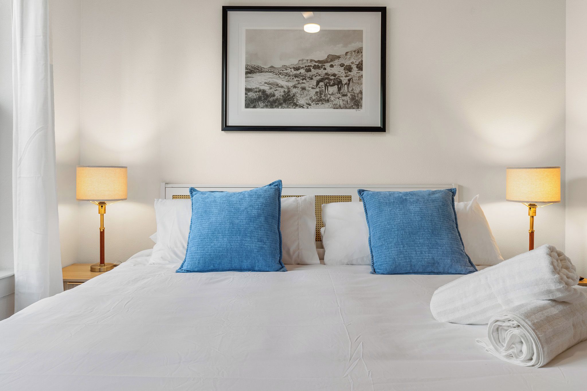 Een hotelkamer met een bed, twee blauwe kussens, een witte deken, een opgerolde handdoek, twee nachtlampjes en een ingelijst schilderij aan de muur.