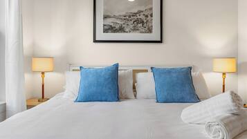 Een hotelkamer met een bed, twee blauwe kussens, een witte deken, een opgerolde handdoek, twee nachtlampjes en een ingelijst schilderij aan de muur.