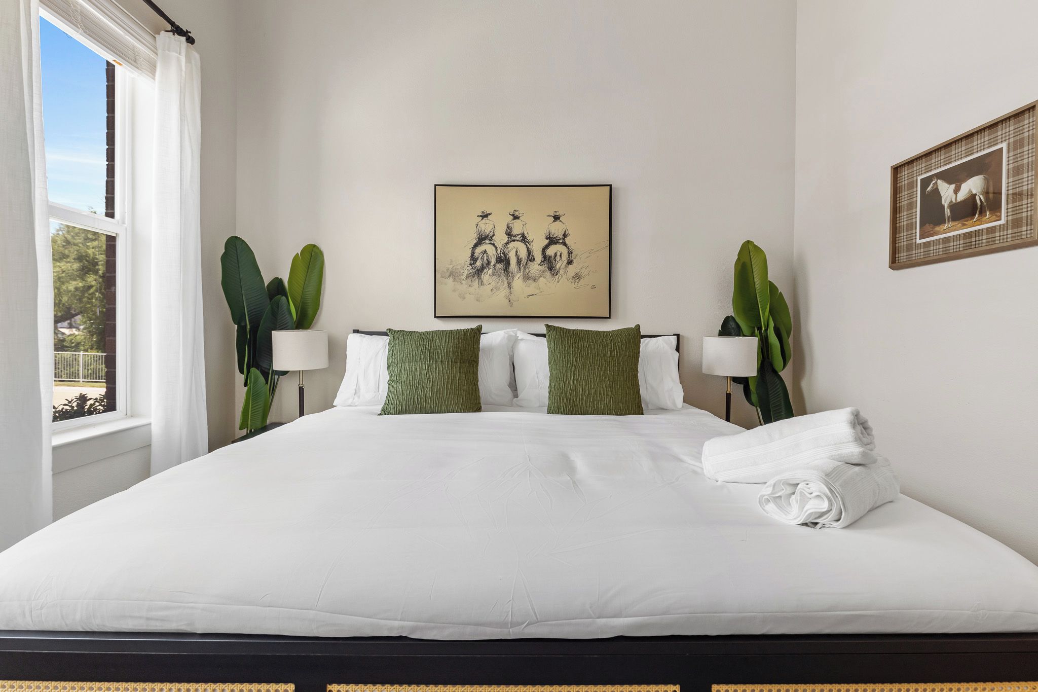 A large bed with white linens, a painting of riders on horses, and two potted plants.