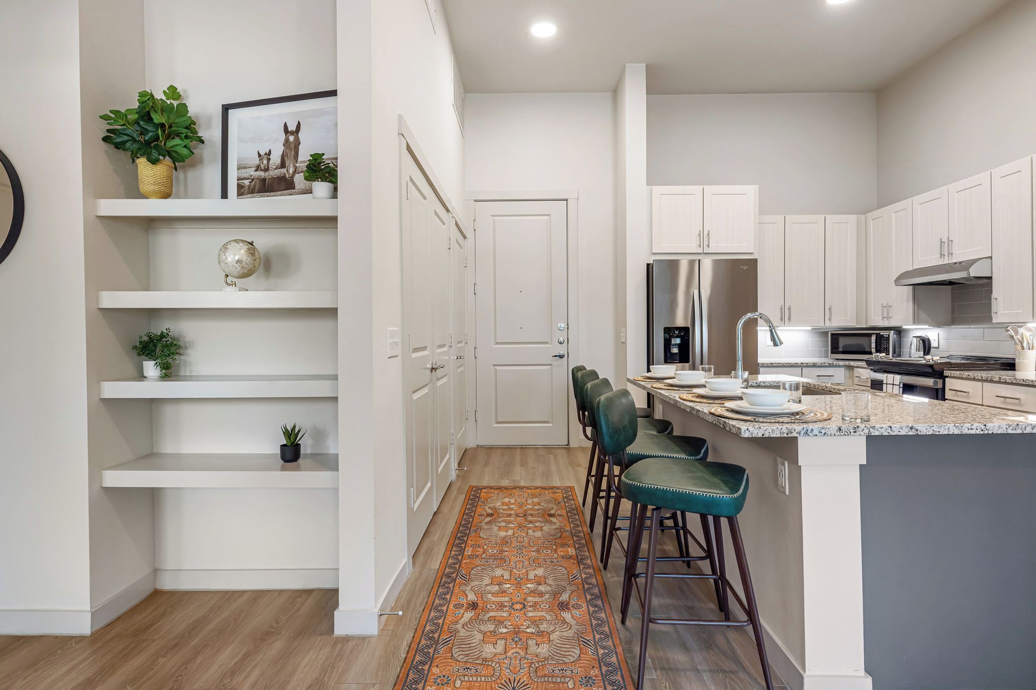 A modern kitchen with white cabinetry, a stainless steel refrigerator, a microwave, and a water dispenser. There are bar stools and a rug in the seating area.