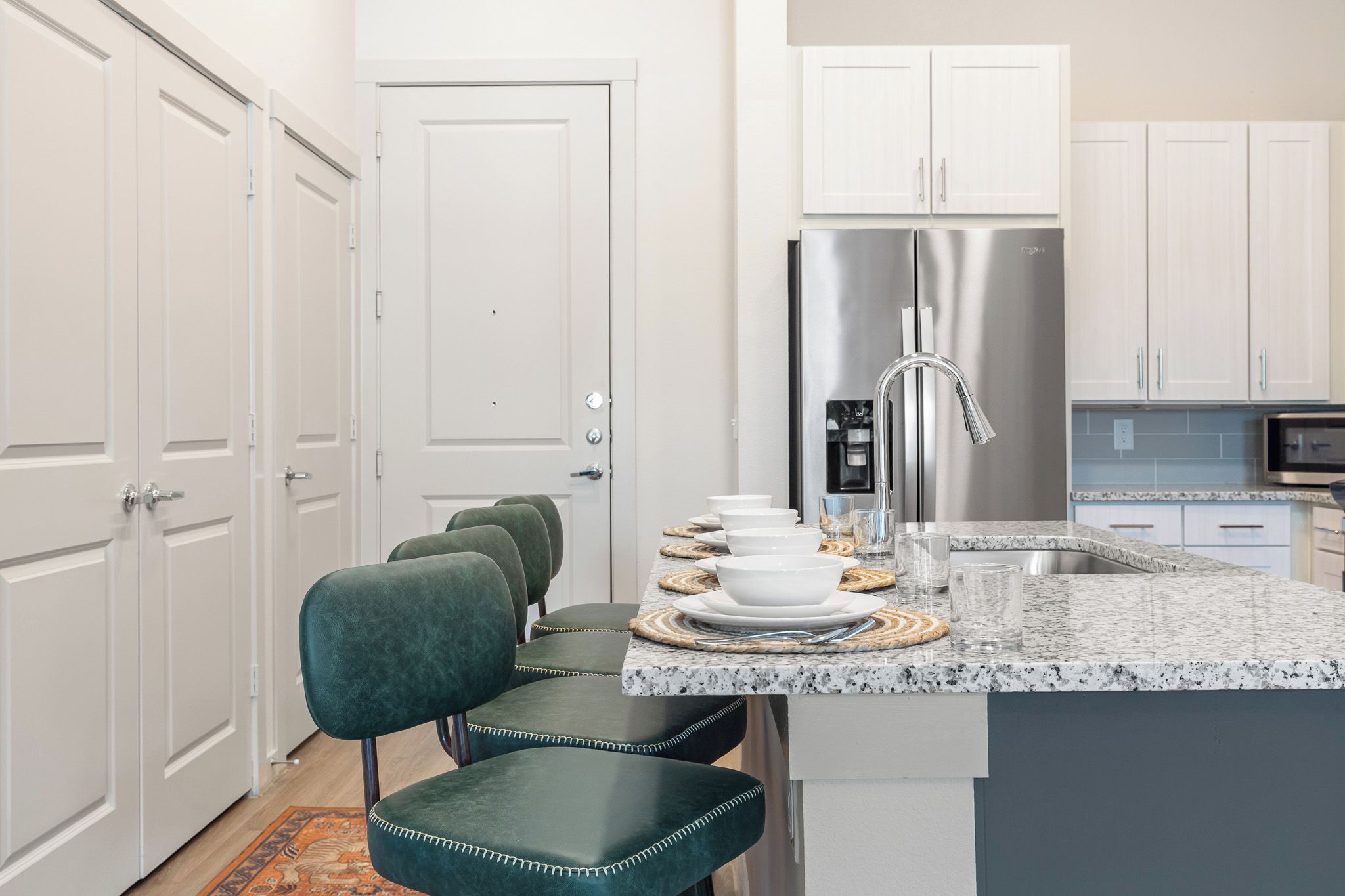 A modern kitchen with white cabinetry, a stainless steel refrigerator, a granite countertop, and a seating area with green upholstered chairs.