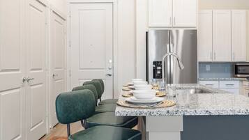 A modern kitchen with white cabinetry, a stainless steel refrigerator, a granite countertop, and a seating area with green upholstered chairs.