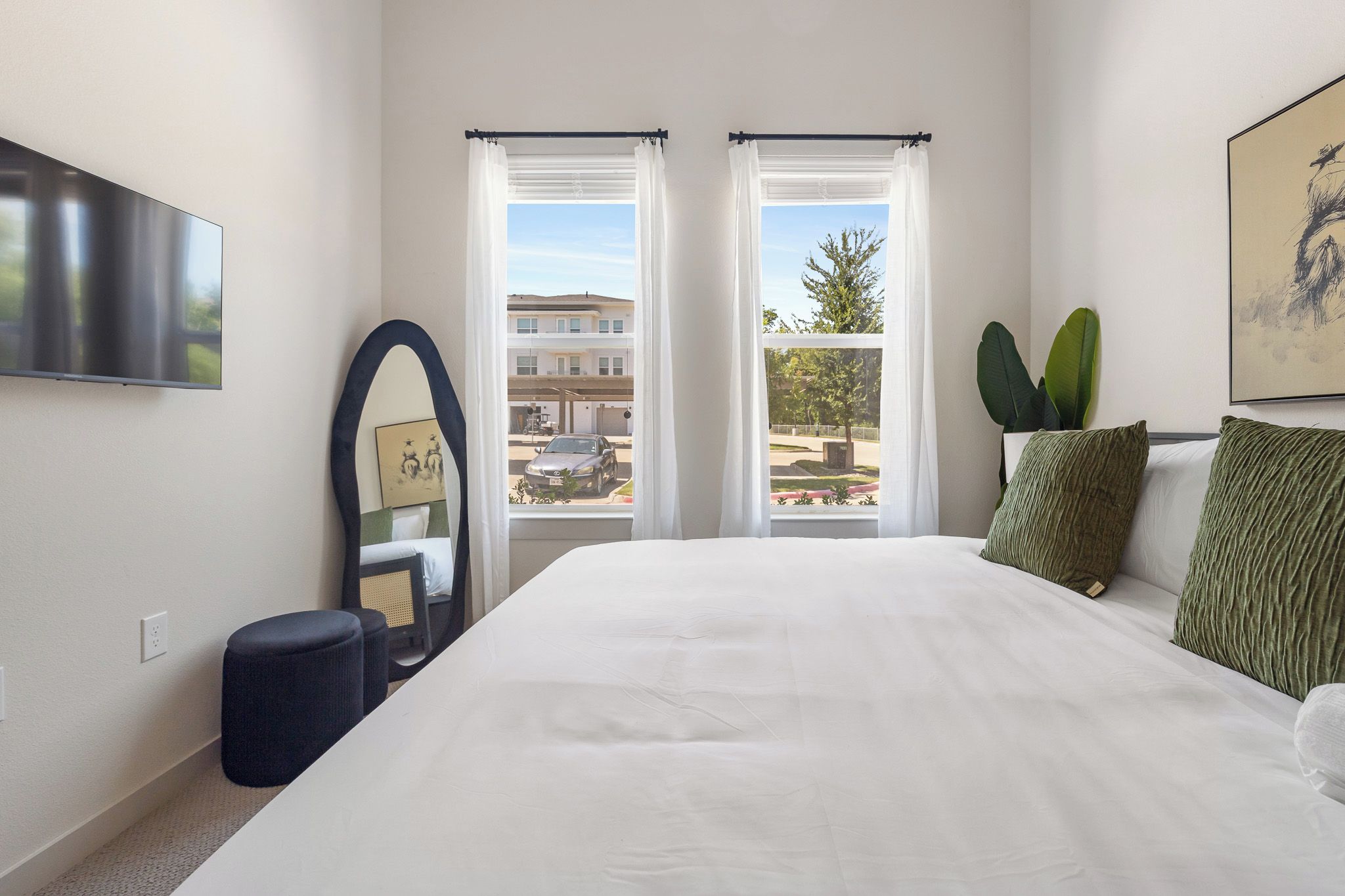 A bedroom with a large bed, two windows with curtains, a mirror, a potted plant, and a painting on the wall.